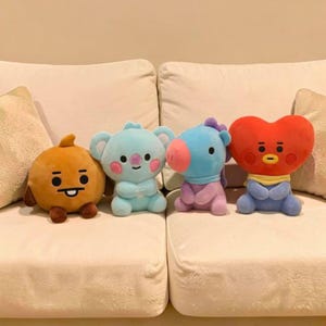 以下が含まれることがあります： 白いソファに座っている、さまざまな色の4つのぬいぐるみキャラクター人形。人形には、丸いオレンジ色のキャラクター、水色のコアラ、青い馬、赤いハート型のキャラクターが含まれています。