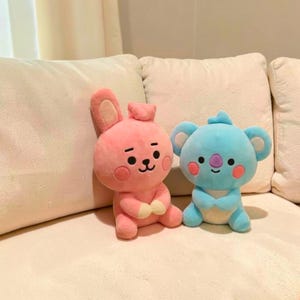以下が含まれることがあります： ピンクのウサギと青いコアラの2つのぬいぐるみ。ウサギは長い耳と小さな尻尾があります。コアラは大きな耳と紫色の鼻を持っています。どちらのおもちゃも頬がピンク色で、白い表面に座っています。