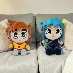 Puede incluir: Dos muñecos de peluche sentados en un sofá gris. Uno tiene cabello castaño, una camisa amarilla con una estrella roja y pantalones azules. El otro tiene cabello azul, un atuendo azul oscuro y una bolsa verde. Ambos muñecos tienen ojos grandes.