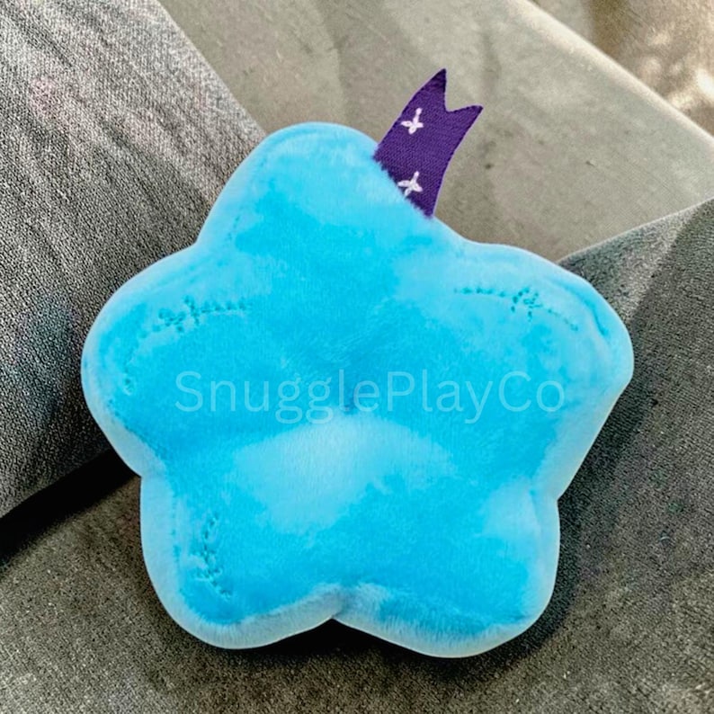 May include: A soft, cloud-shaped toy in shades of blue, with the text "SnugglePlayCo" printed on the front. A purple ribbon with white cross details is attached to the top. The toy is designed for children.