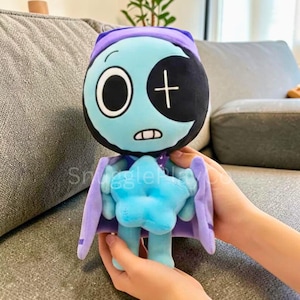 May include: A plush doll with a teal face, a black eye patch, and a purple hood and cape. The doll has a blue star-shaped body and is held by hands. The doll's face has a large white eye and a cross.