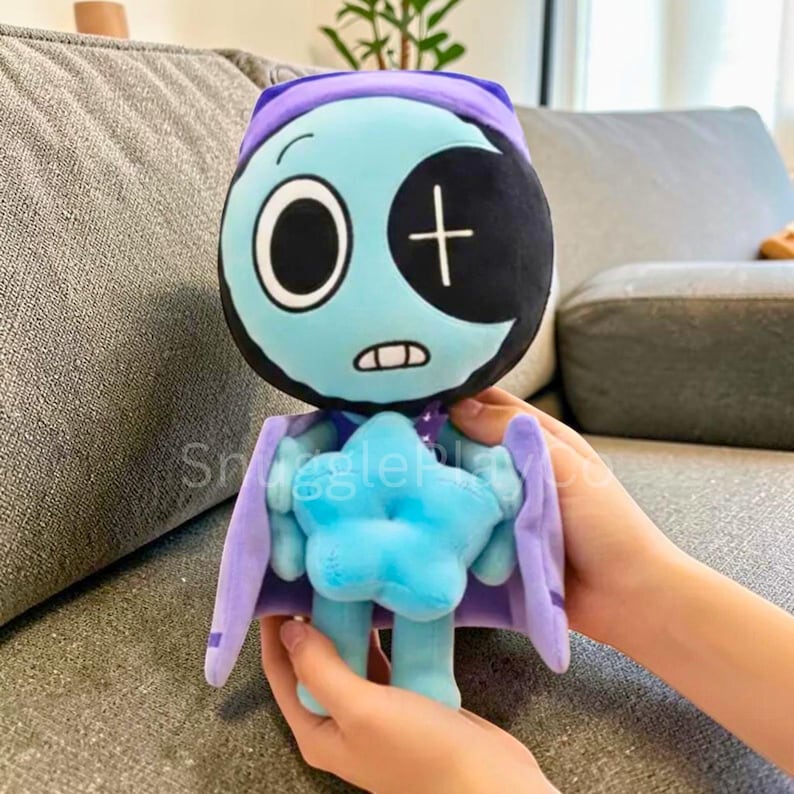 May include: A plush doll with a teal face, a black eye patch, and a purple hood and cape. The doll has a blue star-shaped body and is held by hands. The doll's face has a large white eye and a cross.