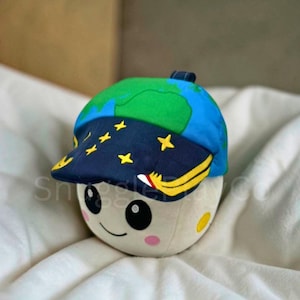 May include: A plush toy shaped like a smiling moon wearing a blue cap with a green Earth design and yellow star accents. The cap's brim features a yellow and white design. The toy has pink cheeks and black eyes.