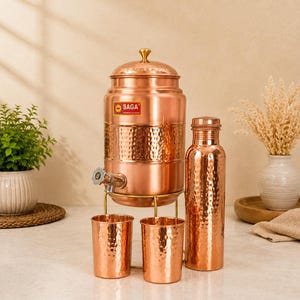 May include: A copper water dispenser with a hammered finish, a metal spigot, and a lid with a gold-tone knob. The set includes two matching hammered copper tumblers and a tall, cylindrical hammered copper water bottle, all on a gold-tone stand.