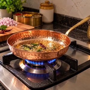 Op de afbeelding: Een ronde, gehamerde koperen koekenpan met een lange messingkleurige steel staat op een brandende gaspit. De pan bevat hete olie met groene chilipepers en gedroogde rode pepers die erin bakken, wat een glinsterend, reflecterend oppervlak creëert.