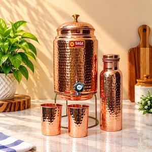 May include: A hammered copper water dispenser with a lid and a black spigot, accompanied by two matching hammered copper tumblers and a tall hammered copper water bottle. The dispenser features a rectangular label with the text SAGA.