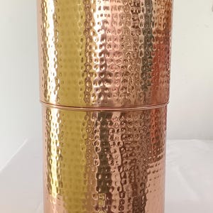 May include: A cylindrical copper water dispenser with a hammered texture, featuring a removable lid with a small knob. The front displays a SAGA Tableware Collection label and a silver-toned spigot at the base, also branded with the SAGA logo.