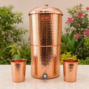 May include: A cylindrical copper water dispenser with a hammered texture, a lid with a small knob, and a spigot at the base. The dispenser features a rectangular label with the text SAGA. Two matching hammered copper tumblers sit on either side.