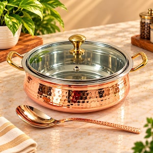 May include: A round, hammered copper cooking pot with a polished stainless steel interior, two gold-toned side handles, and a clear glass lid featuring a gold-toned knob. A matching hammered copper serving spoon with a polished finish is placed in front.