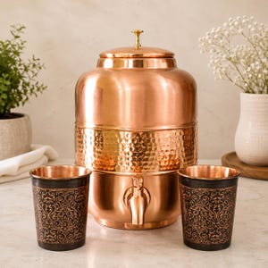 Könnte beinhalten: Ein polierter Wasserspender aus Kupfer mit einem gehämmerten Mittelteil und einem Metallhahn, begleitet von zwei passenden Kupferbechern, die ein dunkles, kompliziertes Blumenmuster auf den Außenflächen jedes Bechers aufweisen.