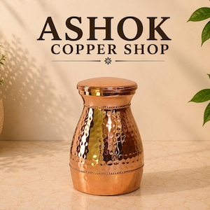 May include: A polished copper vessel with a hammered texture and a smooth, rounded lid. The container features a narrow neck and a wider base, accented by a beaded trim detail. The text ASHOK COPPER SHOP is displayed above the object.