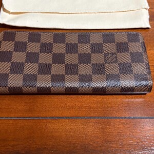 May include: A brown and tan checkered wallet with a red zipper pull. The wallet is rectangular and has a small logo on the front. A cream-colored pouch is in the background.