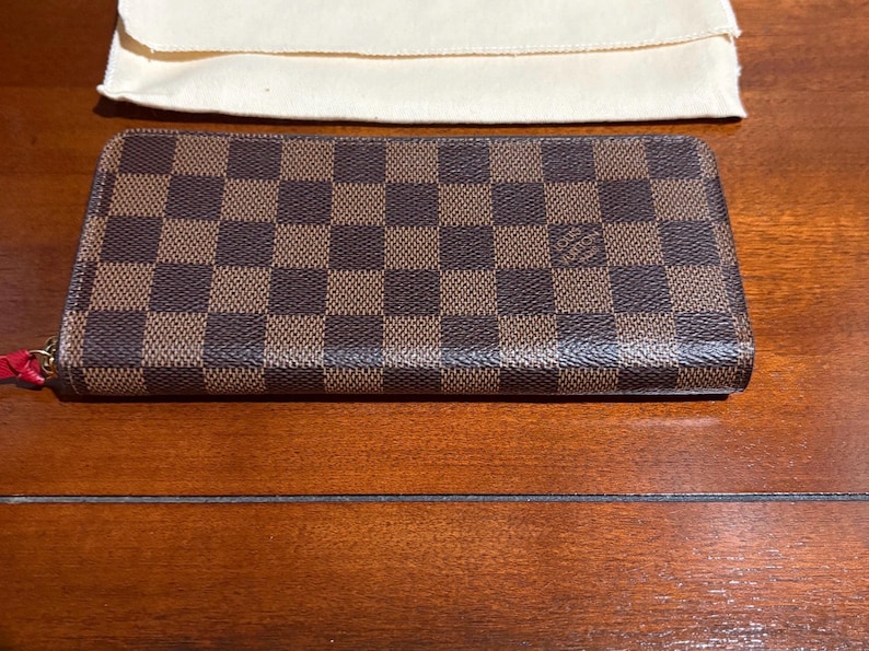 May include: A brown and tan checkered wallet with a red zipper pull. The wallet is rectangular and has a small logo on the front. A cream-colored pouch is in the background.