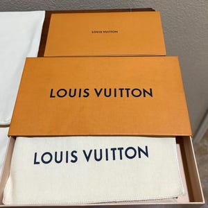 May include: Open orange Louis Vuitton box with the brand name in black. Inside the box is a white cloth bag with the Louis Vuitton logo in navy blue. A smaller orange box with the Louis Vuitton logo is on top.