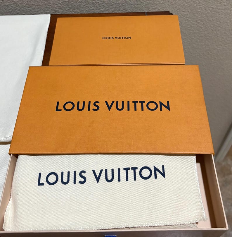 May include: Open orange Louis Vuitton box with the brand name in black. Inside the box is a white cloth bag with the Louis Vuitton logo in navy blue. A smaller orange box with the Louis Vuitton logo is on top.
