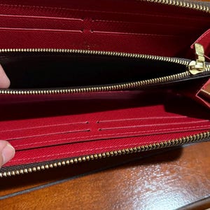 May include: Open, red interior wallet with a brown exterior and a gold zipper. The wallet has multiple card slots and a zippered compartment. The wallet is open, revealing the interior.