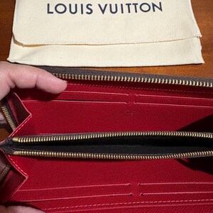 May include: Open brown wallet with a red interior and multiple card slots. The wallet has a gold zipper and is paired with a cream-colored pouch with the text "LOUIS VUITTON" in navy blue.