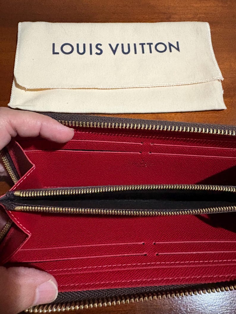 May include: Open brown wallet with a red interior and multiple card slots. The wallet has a gold zipper and is paired with a cream-colored pouch with the text "LOUIS VUITTON" in navy blue.