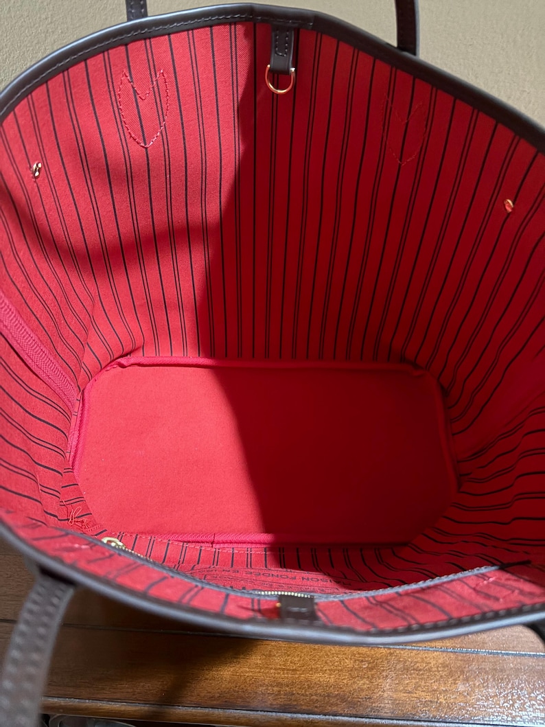 May include: Open view of a brown tote bag with a red interior. The interior features black vertical stripes. The bag has brown handles and gold-colored hardware. The bag's interior has a heart-shaped detail.