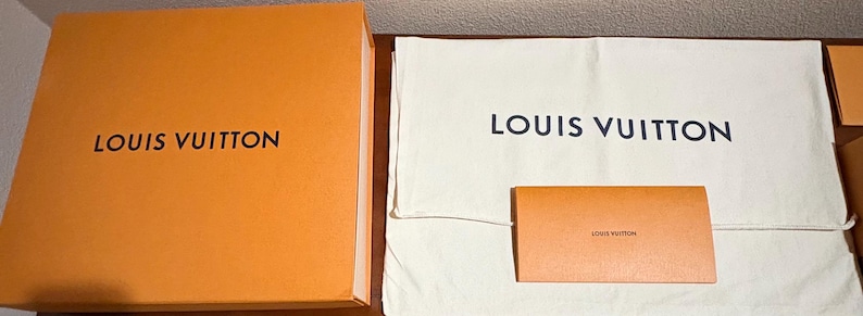 May include: An orange box and a white dust bag, both with the words "LOUIS VUITTON" printed in black. A small orange card with the brand name is also visible.