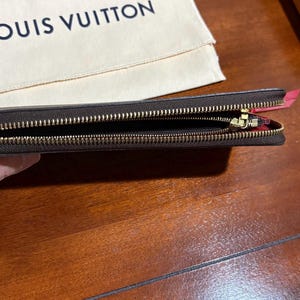 May include: A brown leather wallet with a gold zipper, open to reveal the interior. The wallet is held in a hand, with a beige cloth and the "LOUIS VUITTON" logo in the background. The wallet's interior is red.