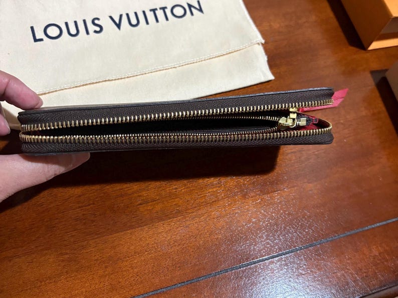 May include: A brown leather wallet with a gold zipper, open to reveal the interior. The wallet is held in a hand, with a beige cloth and the "LOUIS VUITTON" logo in the background. The wallet's interior is red.