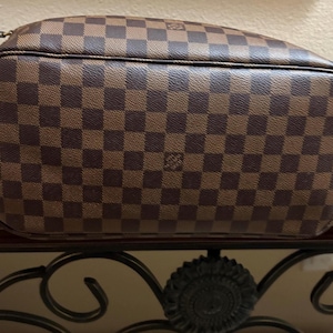 May include: Brown and tan checkered handbag with a rounded shape. The bag features a repeating pattern of squares and a small logo. The bag is made of a durable material and has a shoulder strap.