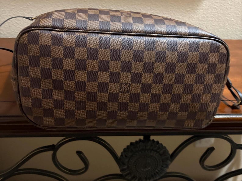 May include: Brown and tan checkered handbag with a rounded shape. The bag features a repeating pattern of squares and a small logo. The bag is made of a durable material and has a shoulder strap.