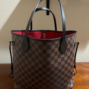 May include: Brown and tan checkered tote bag with dark brown handles and trim. The interior is lined with red fabric. The bag has a structured shape and is sitting on a wooden surface.