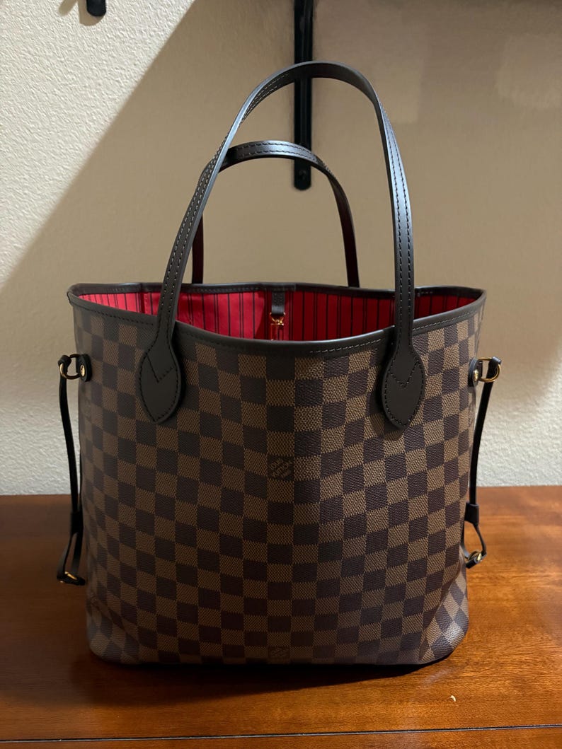 May include: Brown and tan checkered tote bag with dark brown handles and trim. The interior is lined with red fabric. The bag has a structured shape and is sitting on a wooden surface.