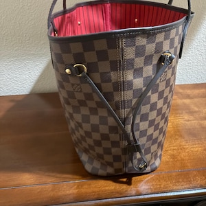 May include: Brown and tan checkered tote bag with a red interior. The bag has two handles and gold-colored hardware. The bag is sitting on a wooden surface.