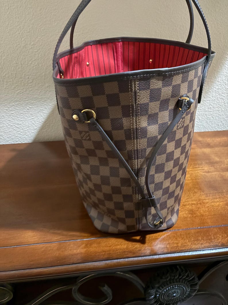 May include: Brown and tan checkered tote bag with a red interior. The bag has two handles and gold-colored hardware. The bag is sitting on a wooden surface.
