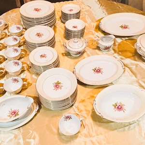 May include: A complete vintage porcelain dinnerware set with a floral rose design. The set includes plates, cups, saucers, serving dishes, a sugar bowl, and a creamer. The dishes are cream-colored with gold trim and a pink rose motif.