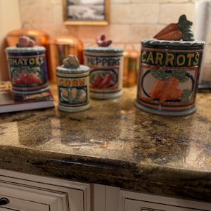 May include: A collection of ceramic kitchen canisters with lids, labeled "Tomatoes," "Corn," "Peppers," and "Carrots." Each canister features a colorful illustration of the corresponding vegetable. The canisters are arranged on a countertop.