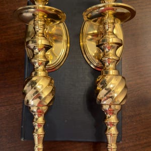 May include: Two ornate, gold-colored wall sconces. Each sconce features a candle cup, a decorative oval backplate, and a twisted, spiral-shaped central column. The sconces are placed on a dark surface.