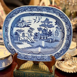 May include: An oval ceramic platter featuring a blue and white Willow pattern. The design depicts a traditional landscape with a pagoda, a bridge, willow trees, and two birds in flight. The border is decorated with a geometric fretwork pattern.
