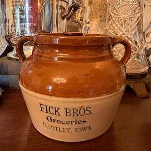 May include: A two-toned stoneware crock with a brown glazed upper section and a tan lower section. It features two side handles and black printed text that reads FICK BROS. Groceries HARTLEY, IOWA. The vessel has a wide mouth and a sturdy base.
