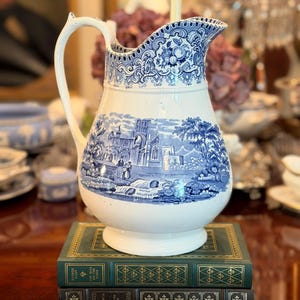 May include: A white ceramic pitcher featuring a blue transferware pattern of a landscape with buildings and figures. The rim has a decorative pierced lattice design. The pitcher is resting on two stacked green and black hardcover books.