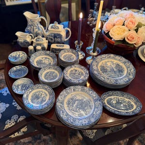 May include: A collection of blue and white porcelain tableware, including plates, saucers, a teapot, and serving dishes, arranged on a dark wooden table. The plates feature a detailed landscape design. A bouquet of pink and yellow roses and lit candles are also on the table.