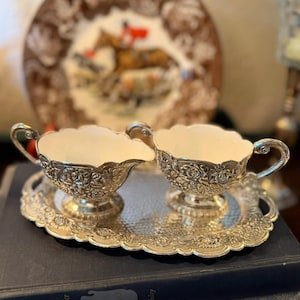 May include: A silver-colored creamer and sugar bowl set with floral detailing, resting on a matching tray. The set is placed on top of a stack of books. A decorative plate with a horse and rider is in the background.