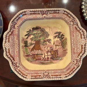 May include: A square ceramic bowl with scalloped edges featuring a cream-colored center with a transferware scene of a horse-drawn cart in a landscape. The wide border is decorated with a dark red and white intricate scrollwork pattern.