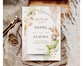 以下が含まれることがあります： 「Save The Date」の文字と「Daniel」と「Claudia」の名前が入った結婚式の招待状。ニュートラルな色調の花柄とパンパスグラスのアクセント、アーチ型、エレガントな筆記体デザインです。