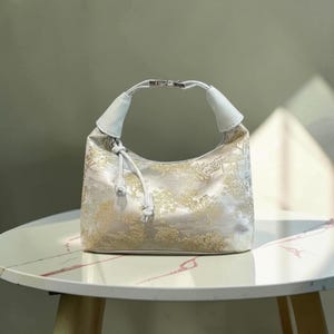 May include: A cream-colored handbag with a gold floral pattern. The bag has a curved shape with a white handle and drawstring closure. The bag is sitting on a white and gray table.