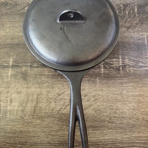 May include: A black cast iron skillet with a matching circular lid. The lid features a small, arched handle with a central hole. The skillet has a long, slender handle with an elongated opening in the center, extending from the circular base.