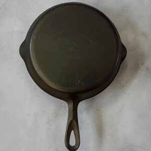 May include: A round, dark gray cast iron skillet viewed from the underside. The pan features a smooth, circular base with a prominent number 5 embossed in the center. It includes a single handle with a teardrop-shaped hole at the end for hanging.
