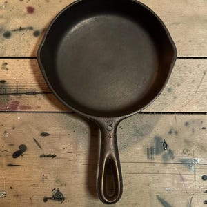 May include: A cast iron skillet with a single handle and a rounded cooking surface. The skillet is dark brown and appears well-seasoned. The handle has a hole for hanging and the number "3" is visible on the handle.