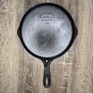 May include: A black cast iron skillet with a round base and a single handle. The underside features the embossed text WAGNER WARE SIDNEY -O- and the number 1056. The surface shows signs of use with a dark, seasoned finish and a smooth, circular design.