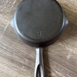 May include: A round black cast iron skillet featuring a single long handle with a teardrop-shaped hole at the end. The underside of the pan displays the number 3 embossed in the center, along with two small pouring spouts on opposite sides.