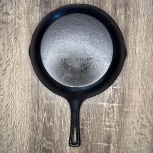 May include: A black cast iron skillet with a long handle featuring a central cutout. The underside is embossed with the text CHEF SKILLET, 8 INCH, MADE IN USA, and a single X mark. The surface has a textured, dark metallic finish.