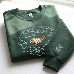 K&ouml;nnte beinhalten: Ein waldgr&uuml;ner Rundhals-Pullover mit einer Stickerei eines beigen und braunen fliegenden Bisons, umgeben von hellblau gestickten Wolken. Der Text YIP YIP ist in Hellblau auf dem rechten &Auml;rmelb&uuml;ndchen des Kleidungsst&uuml;cks eingestickt.